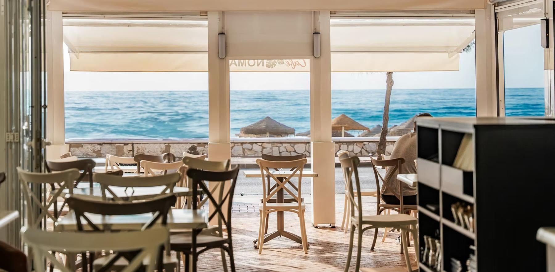 The dining room open onto the terrace: straw parasols, two guests at a table and the Mediterranean.